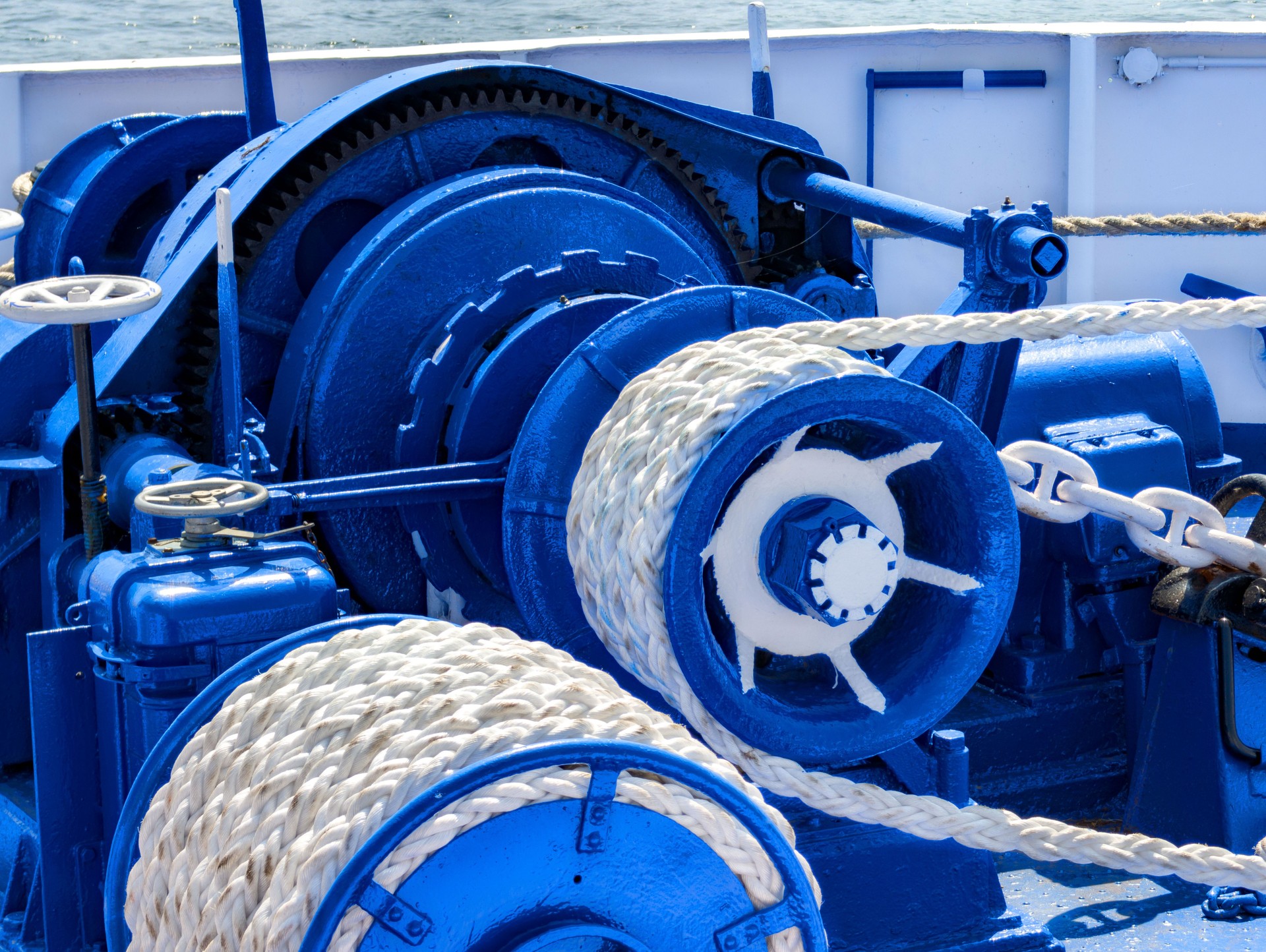 Detail of marine equipment showcasing a blue winch and rope on a vessel, under bright sunlight by the water, capturing the intricacies of nautical machinery